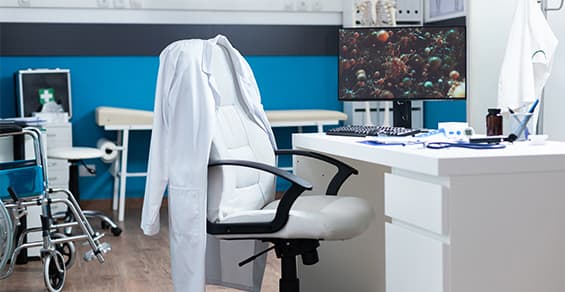 Clinic consultation room with a white coat over the desk chair, a computer desk, an examination bed and a wheelchair